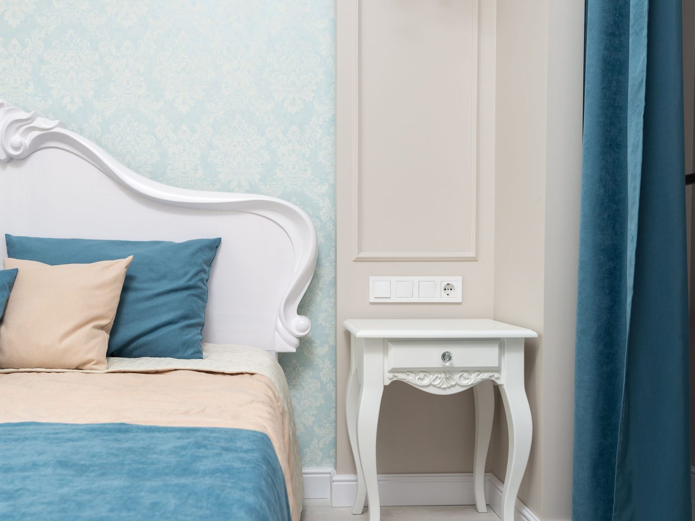Bedroom with pale blue damask wallpaper meeting a cream panelled wall, a white carved headboard and a small white nightstand