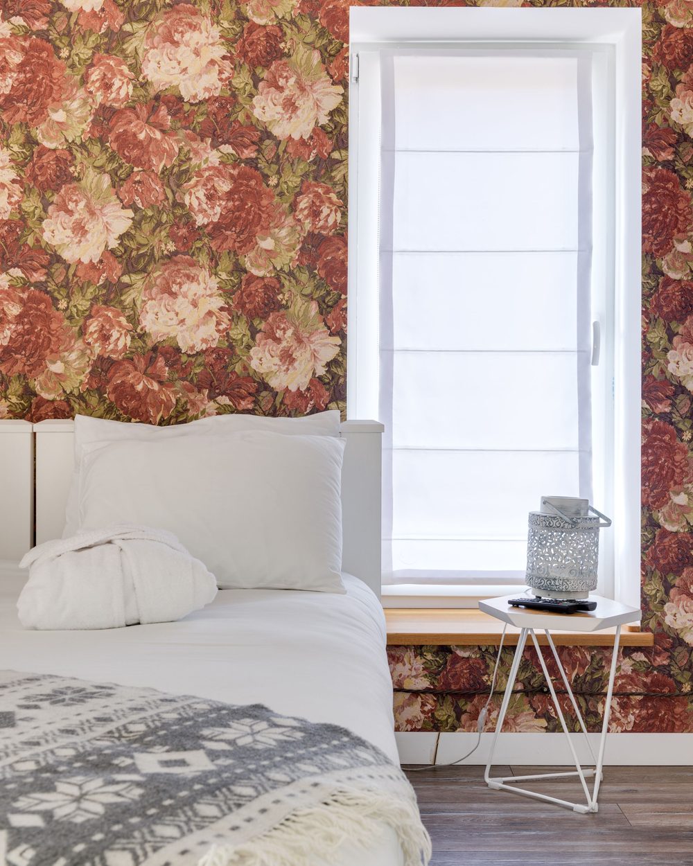 Guest bedroom with a deep red and cream large floral wallpaper cut neatly around a window, white bedding and a wire side table