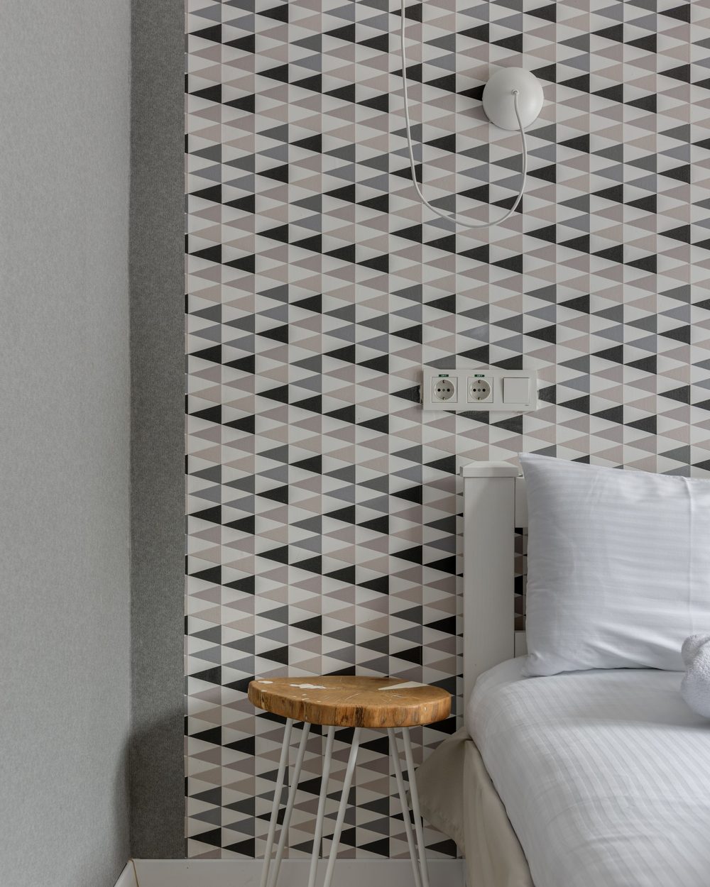 Black, grey and white triangle-pattern accent wall beside a white bed and a live-edge wooden stool