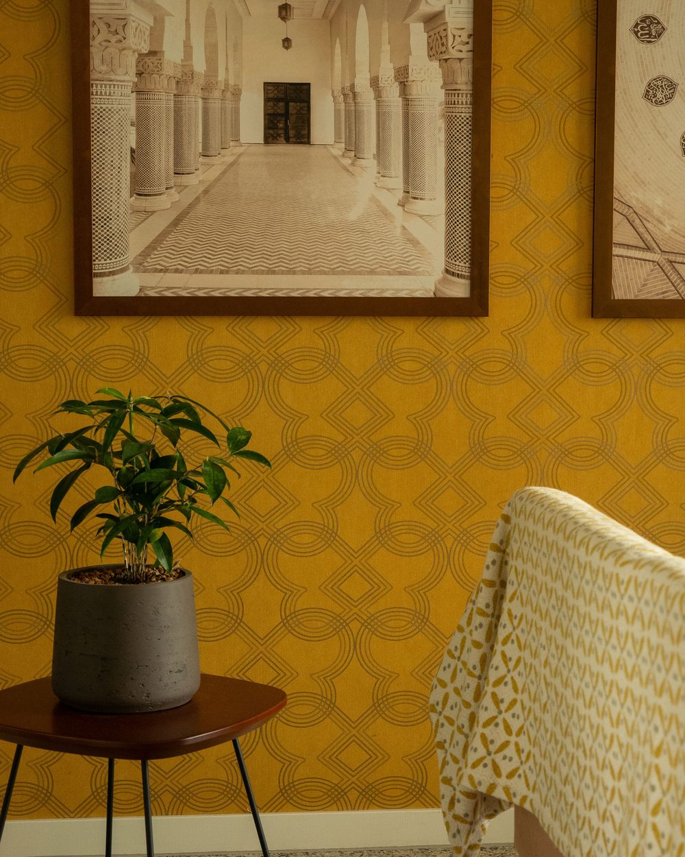Ochre trellis-pattern wallpaper with a large framed print, a potted plant on a side table and the arm of a sofa