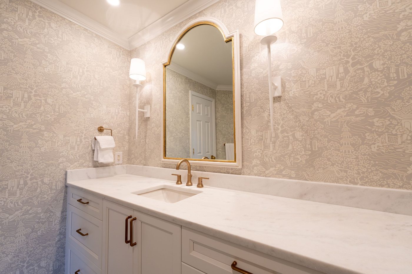 Bathroom with cream toile wallpaper, crown moulding, a brass arched mirror, twin sconces and a white vanity with marble top