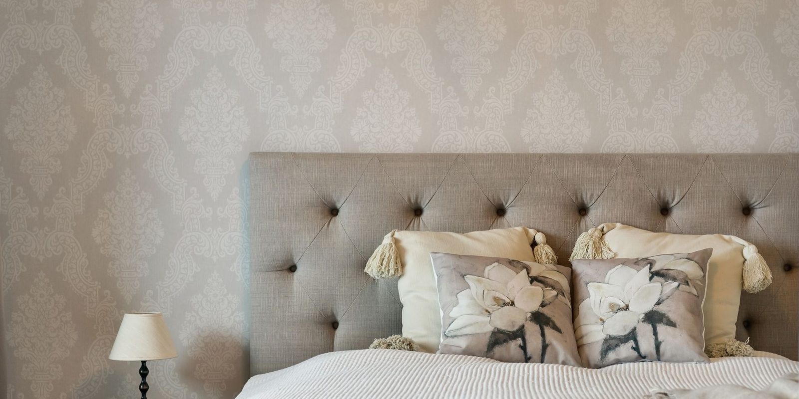 Bedroom with a taupe damask wallpaper behind a tufted linen headboard, cream cushions and a small table lamp