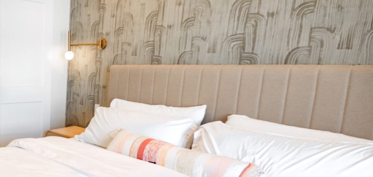 Warm grey brushstroke wallpaper behind a channel-tufted linen headboard with white bedding and a brass sconce