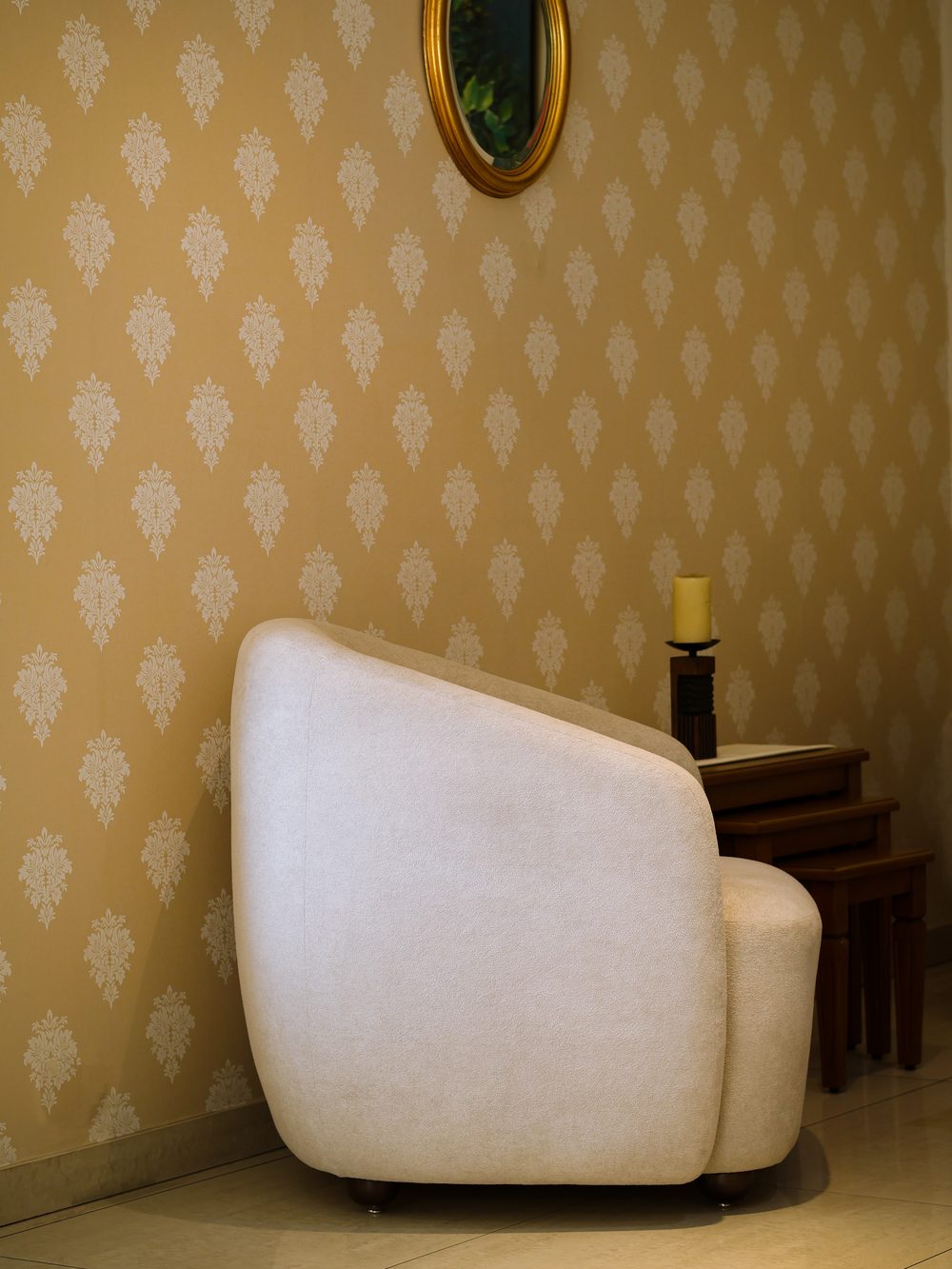 Living room reading corner with an ochre damask wallpaper, a cream curved armchair and a small oval mirror