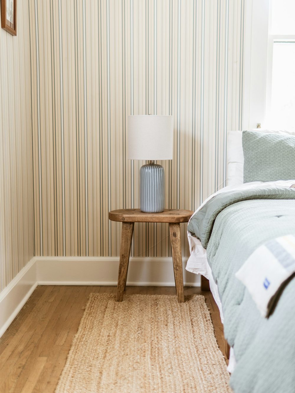 Bedroom corner with fine cream pinstripe wallpaper, a wooden stool with a table lamp and a woven rug