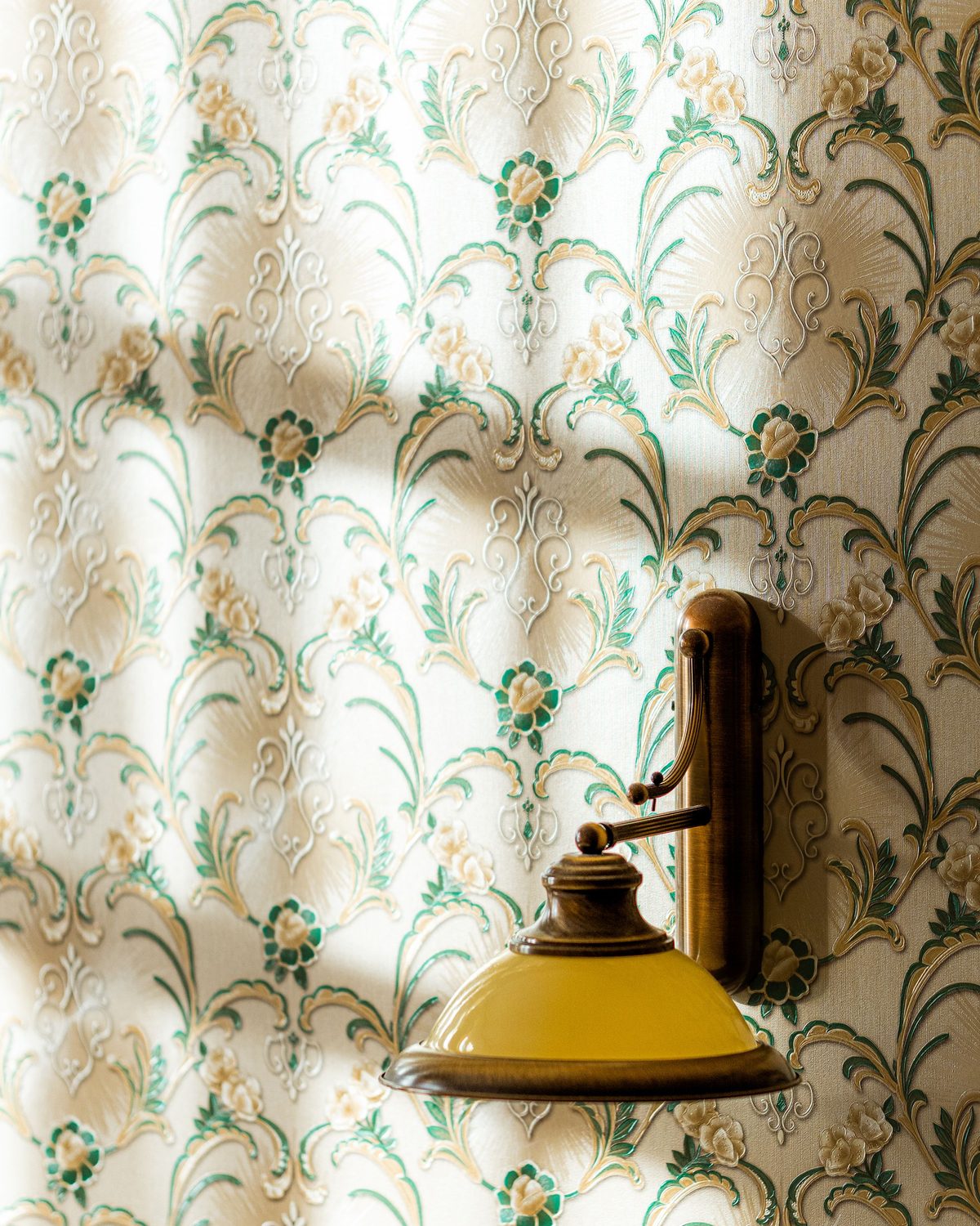 Sunlight falling across a cream and green scrolling floral wallpaper with a small brass wall lamp